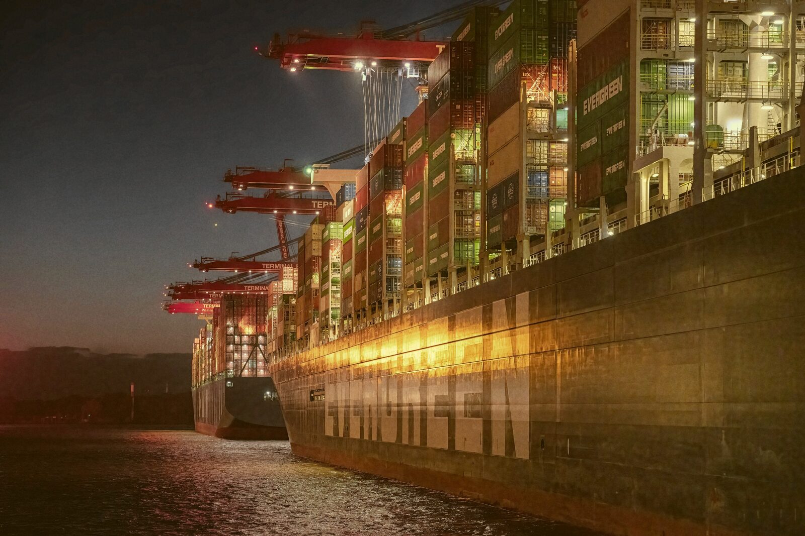 A container ship loaded with cargo docked beside gantry cranes at dusk.