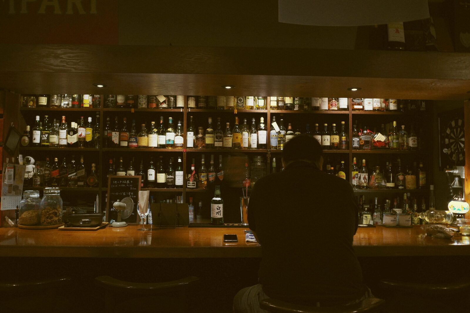 A silhouetted figure seated at a long bar backed by shelves of bottles.