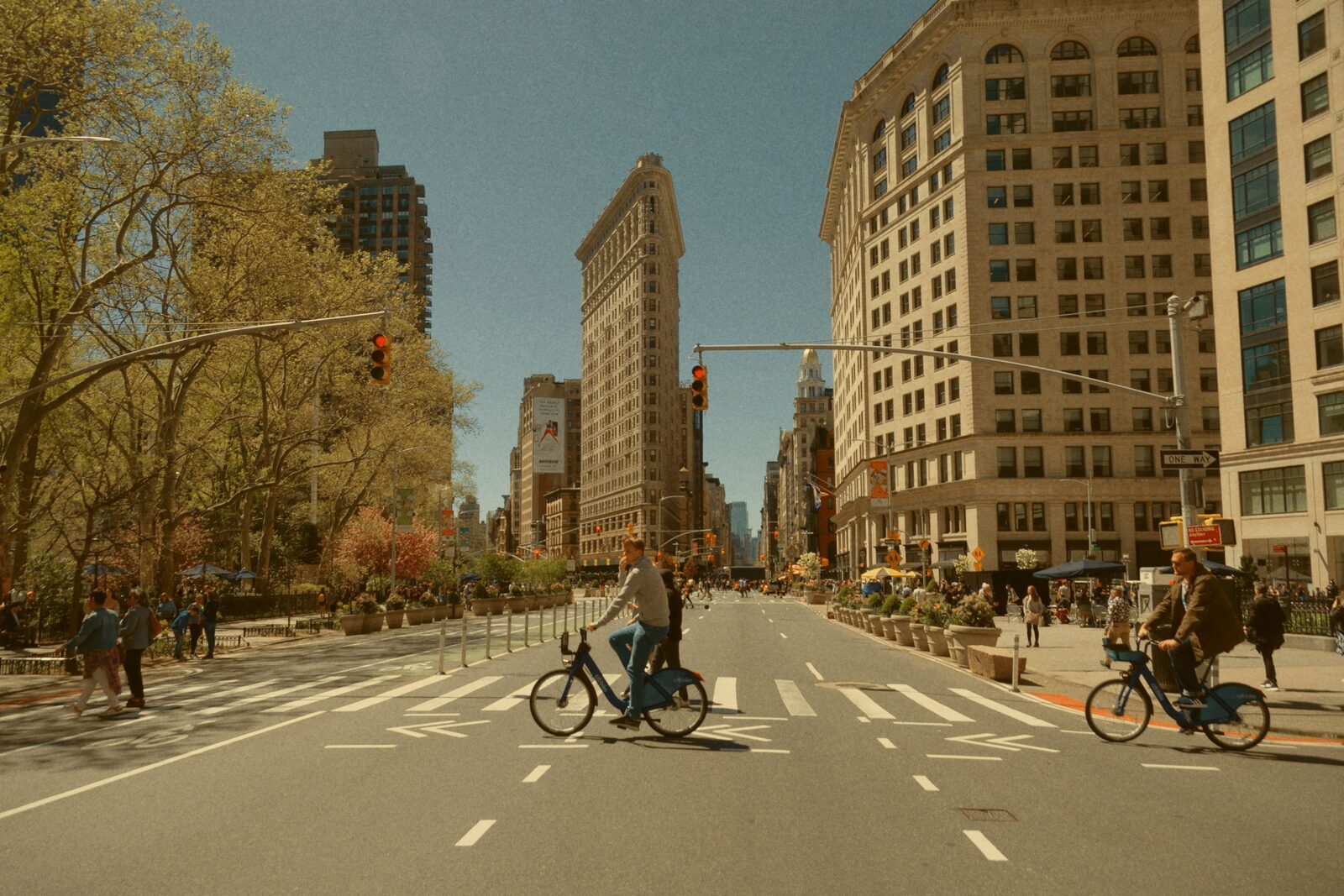 Cyclists cross a wide city avenue near a triangular high-rise building.