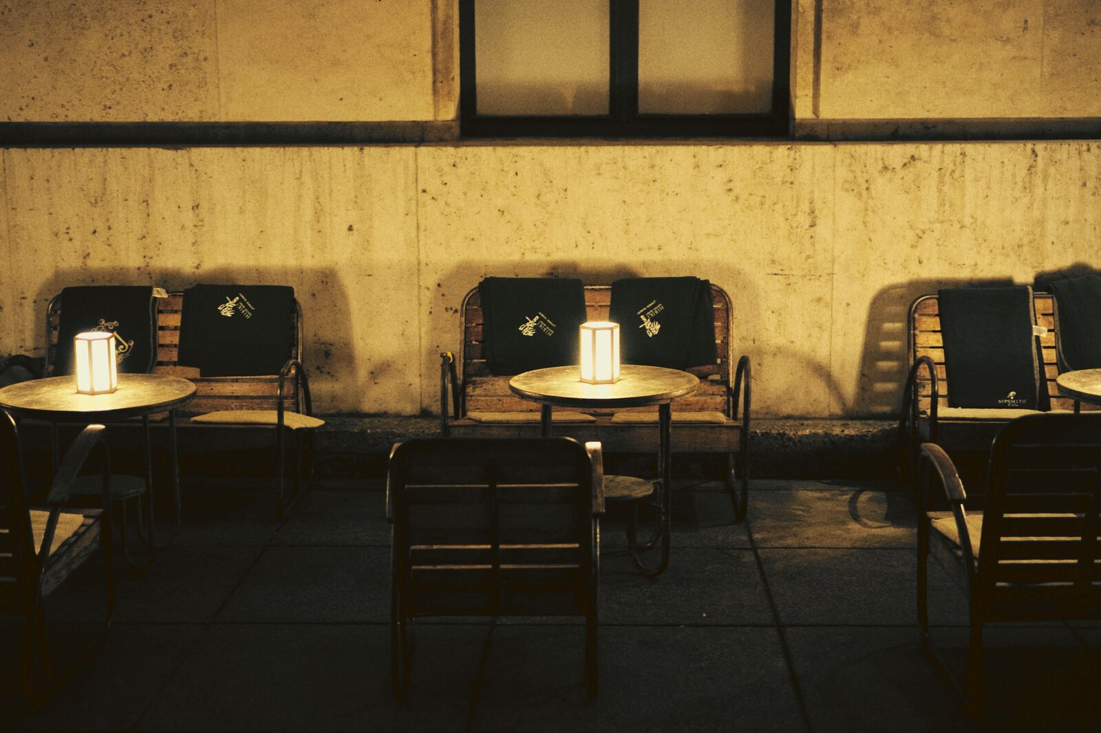Empty café tables and chairs lit by candlelight against a wall at night.