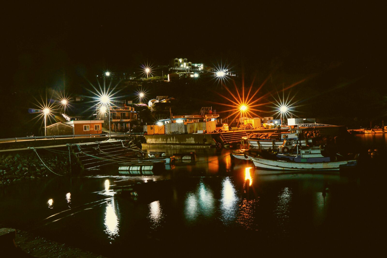 Small boats moored at a harbor dock lit by starburst lights at night.