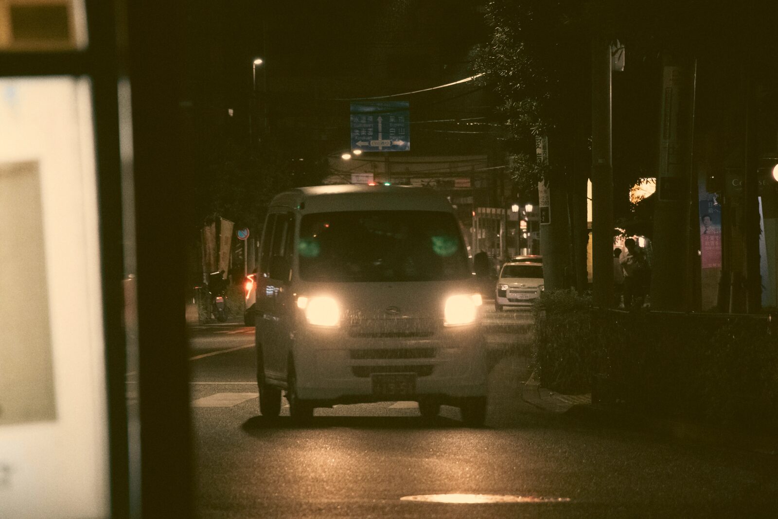 A small delivery van driving down a city street at night, headlights lit.