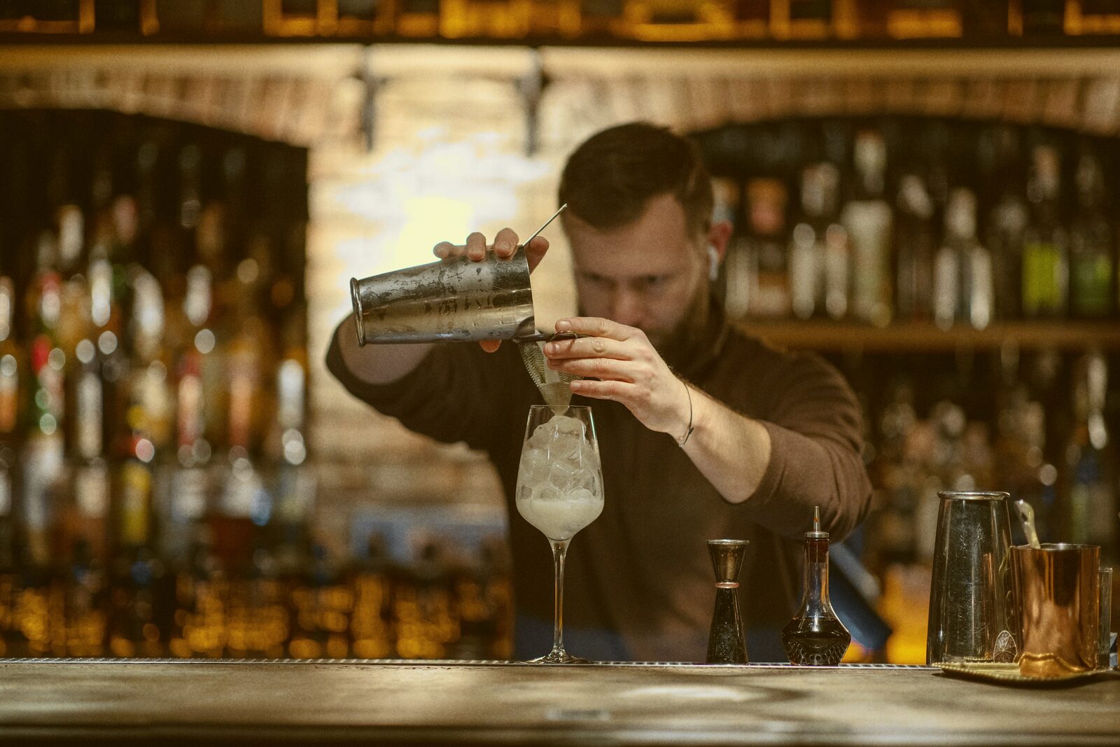 A bartender pours a frothy cocktail into a glass in a dim bar.